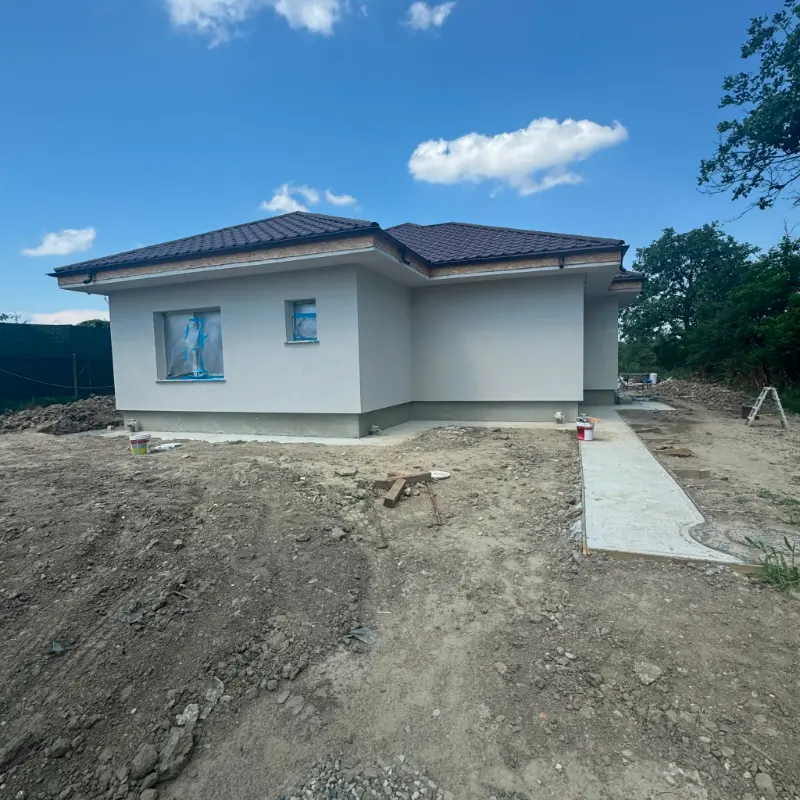 Casă parter cu tencuială decorativă gri, acoperiș în patru ape din țiglă metalică maro și trotuar de gardă turnat